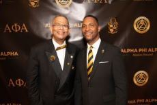 Two men in black suits. One bow tie and one with black/gold regular tie. Black and gold backdrop.