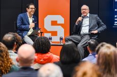 Coach Boeheim and gentleman sitting with microphones with pull up banner-white block S on orange background.