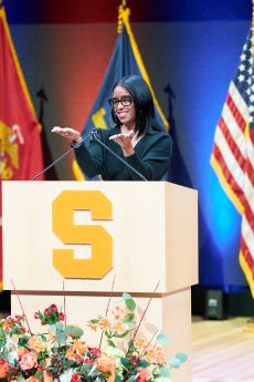 Alumni wearing glasses holding out her hands with orange block S on white podium and partial view of American flag.