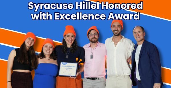 The staff of Syracuse Hillel stand proudly with Hillel International CEO, Adam Lehman as he presents them with an excellence award. They are standing on a blue and orange striped background and they're all wearing matching orange hats. The banner reads, Syracuse Hillel Honored with Excellence Award